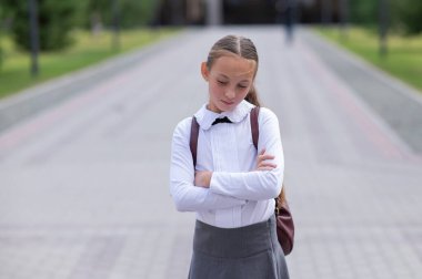 Üniformalı, sırt çantalı, kollarını göğsüne kavuşturmuş üzgün, beyaz bir kız okuldan sonra dışarıda duruyor.