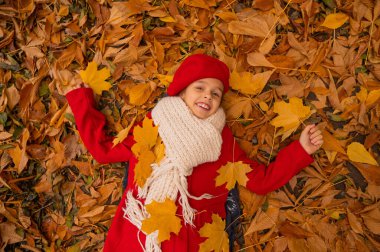 Kırmızı paltolu ve bereli beyaz bir kızın yukarıdan görünüşü sarı yaprakların üzerinde yatıyor. Sonbaharda parkta yürüyüş