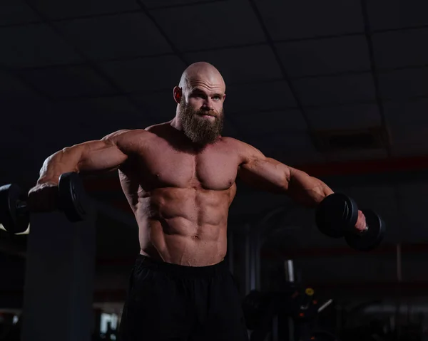 A muscular shirtless man lifts dumbbells to the sides with dumbbells in the gym