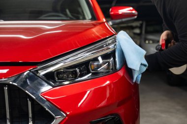 Bir adam mikrofiber bez ve sprey kullanarak kırmızı bir arabanın farlarını yıkıyor.