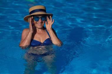 Havuzda mavi gözlüklü ve şapkalı yaşlı bir kadının portresi yüzüyor.