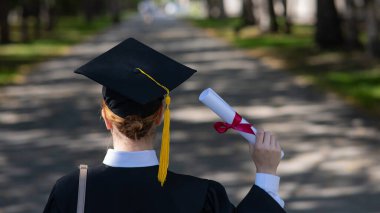 Rear view of a caucasian woman in a graduate gown holding a diploma outdoors