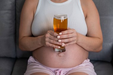 Kanepede otururken elinde bir bardak bira tutan hamile bir kadının göbeğine yakın plan.