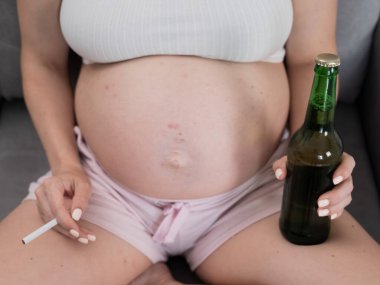 Suratsız hamile kadın kanepede otururken bira içiyor.