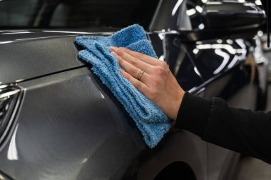 Bir adam gri bir arabanın yüzeyini mikrofiber bir bezle siliyor.