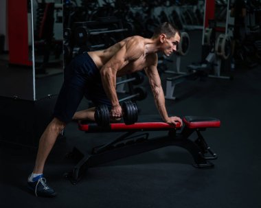 Gömleksiz bir adam spor salonunda dambıl sırasına dizer.