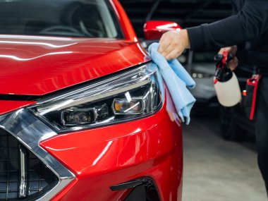Bir adam mikrofiber bez ve sprey kullanarak kırmızı bir arabanın farlarını yıkıyor.