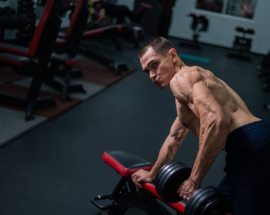 Gömleksiz bir adam spor salonunda dambıl sırasına dizer.