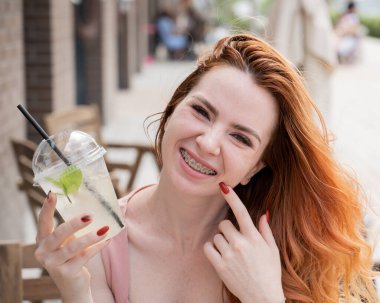Diş teli takan güzel, kızıl saçlı bir kadın yazın dışarıda soğuk limonata içiyor. Çilli, gülümseyen bir kızın portresi.