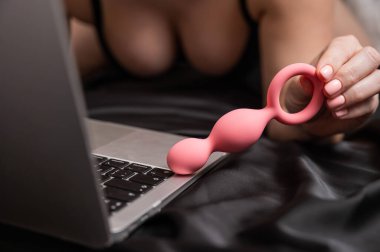 Laptopun yanında pembe anal boncuklar tutan kadın siyah çarşafın üzerinde yatıyordu.