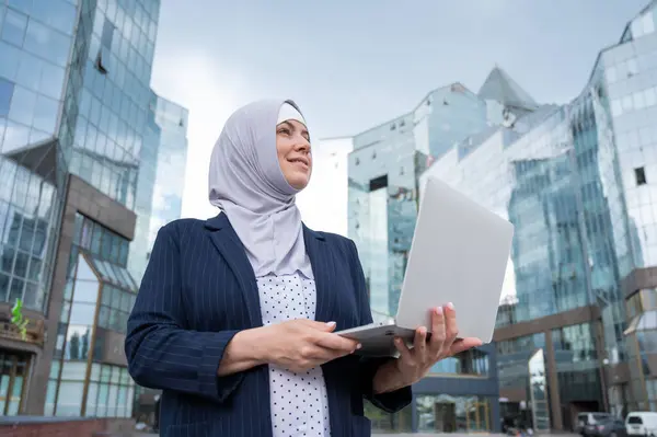 Thoughtful business woman in hijab and suit is holding a laptop outdoors