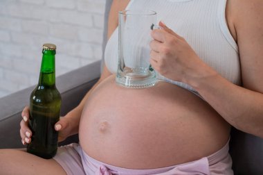 Kanepede oturan, elinde bira şişesi ve bardakla yüzü olmayan hamile kadın.