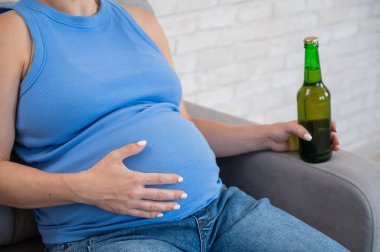 Suratsız hamile kadın kanepede otururken bira içiyor.