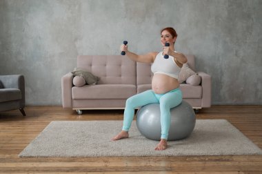 Hamile kadın evde spor yaparken dambıllarla egzersiz yapıyor.