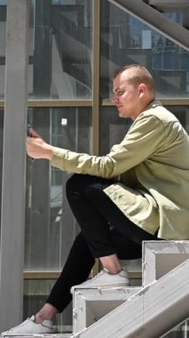 Genç adam merdivenlerde oturup dışarıdaki akıllı telefondan işaret diliyle konuşuyor. Dikey video
