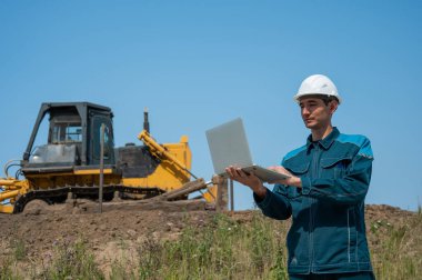 İnşaat alanında elinde dizüstü bilgisayar tutan beyaz erkek müteahhit.