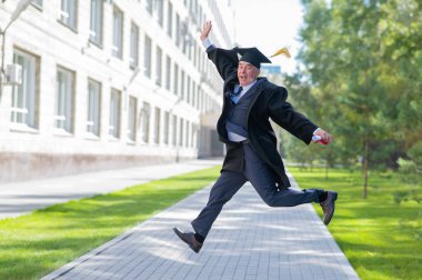 Mezuniyet elbisesi giymiş, dışarı atlayan ve diploması olan yaşlı mutlu bir adam.