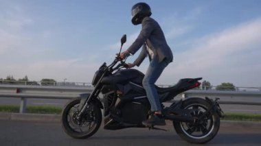Kafkasyalı adam, elektrikli motosiklet sürerken ayağa kalkıp bacaklarını doğrultuyor.