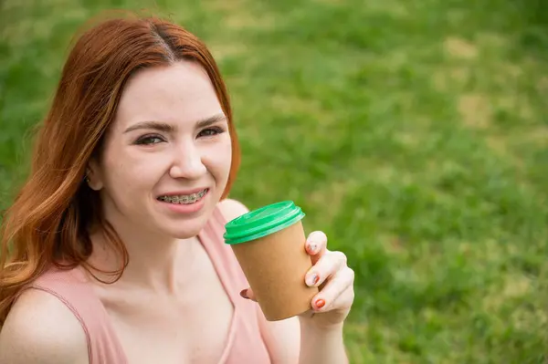 Dişlerinde diş teli olan güzel genç bir kadın çimenlerde otururken karton bir bardaktan içiyor.