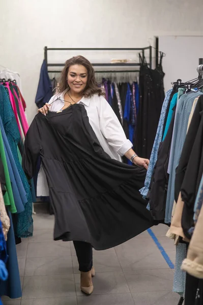 A fat woman in a plus size store tries on a black evening dress. Vertical photo