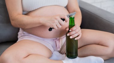 Kanepede oturan ve bira şişesini açan yüzü olmayan hamile kadın.