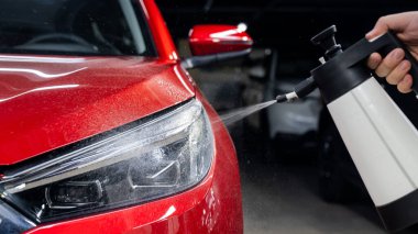 Bir adam kırmızı bir arabanın farlarını spreyle yıkıyor.