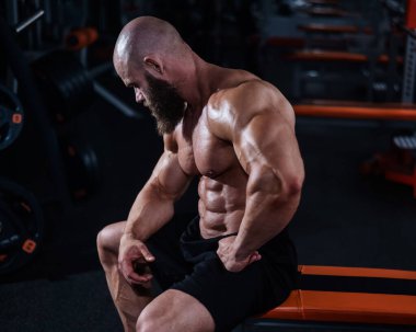 Kısa şortlu, kaslı, kel bir adam antrenmandan sonra bankta dinleniyor. Vücut geliştirici spor salonunda vücudunu gösteriyor.