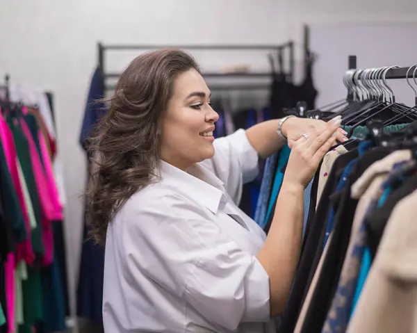 A smiling fat woman in a plus size store chooses clothes while sorting through the hangers