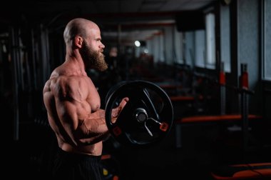 Beyaz, kel, üstsüz bir adam spor salonunda halterle egzersiz yapıyor. Ağırlıklarla bibik bukleler