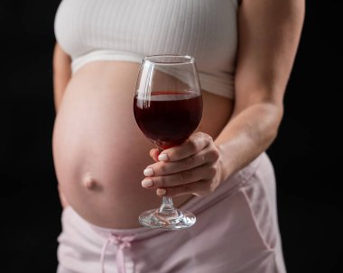 Kanepede otururken elinde bir bardak kırmızı şarap tutan hamile bir kadının göbeğine yakın plan.