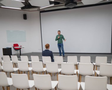 Kızıl saçlı beyaz bir iş kadını boş bir konferans odasının ön sırasında oturuyor. Sakallı adam ders veriyor.