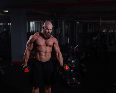 Spor salonunda dambıllarla egzersiz yapan kaslı üstsüz bir adam.