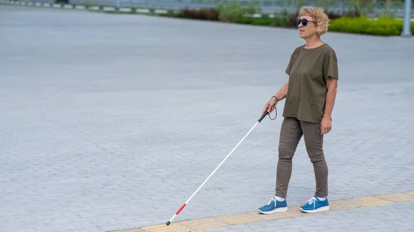 An elderly blind woman walks with a cane along a tactile tile