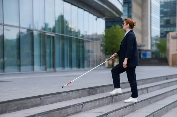 Blind business woman in glasses and with a cane climbs the stairs to the business center