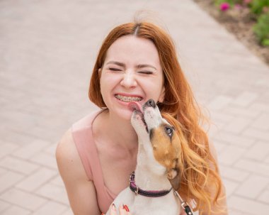 Köpek Jack Russell Terrier sahibinin yüzünü açık havada yalıyor. Dişlerinde diş teli olan kız.