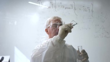 Koruyucu takım elbiseli yaşlı bir beyaz erkek kimyager bir test tüpü tutuyor ve bardağın üzerine bir formül yazıyor.