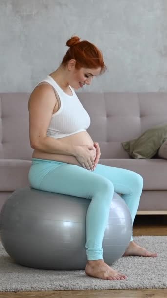 赤毛の妊婦が自宅でフィットボールに座っている間に腹をストロッキング — ストック動画