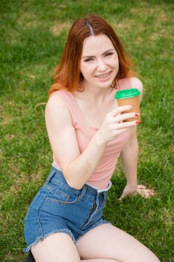Dişlerinde diş teli olan genç ve güzel bir kadın çimlerin üzerinde otururken karton bir bardaktan su içiyor. Dikey fotoğraf