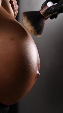 Usta, bronzlaştırma işleminden sonra hamile bir kadının cildine ışıltıyla pudra sürer. Karnına yakın çekim yap. Dikey fotoğraf