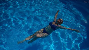 Güneş gözlüklü yaşlı bir kadın havuzda sırtüstü yüzüyor. Emeklilikte tatil