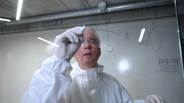 Koruyucu takım elbiseli yaşlı bir beyaz erkek kimyager cama yazı yazıyor.