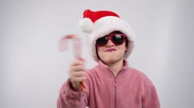 Beyaz arka planda şeker kamışını gösteren, Noel Baba şapkalı ve güneş gözlüklü beyaz bir kızın portresi.