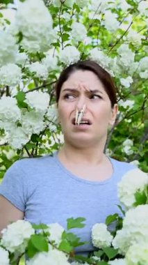 Mutsuz bir kadın burnunda çicek tokasıyla çiçek açan bir ağacın yanında duruyor. Alerji. Dikey video