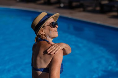 Hasır şapkalı, güneş gözlüklü ve mayolu yaşlı bir kadının portresi havuz kenarında dinlenirken tenine güneş kremi sürüyor.