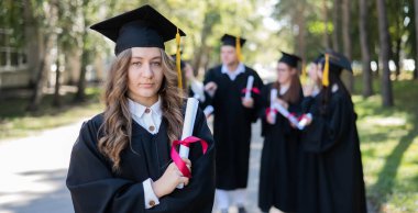 Mezuniyet elbisesi giymiş bir grup mutlu öğrenci dışarıda. Ön planda elinde diploması olan genç bir kız.