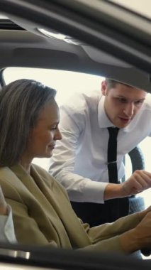 Beyaz saçlı, beyaz bir kadın bir arabanın direksiyonunda oturuyor. Araba galerisi satıcısı olgun bayana danışıyor. Dikey video