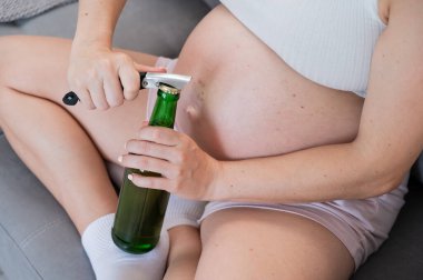 Kanepede oturan ve bira şişesini açan yüzü olmayan hamile kadın.