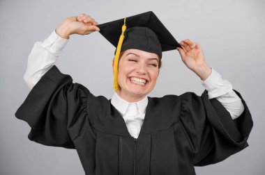 Mezuniyet elbisesi içinde gülümseyen kadın beyaz arkaplanda püsküllü şapka taşıyor.