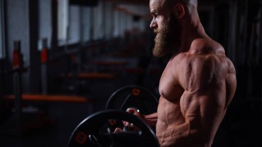 Beyaz, kel, üstsüz bir adam spor salonunda halterle egzersiz yapıyor. Ağırlıklarla bibik bukleler