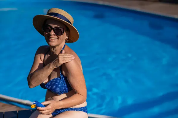 Portrait of an old woman in a straw hat, sunglasses and a swimsuit applying sunscreen to her skin while relaxing by the pool